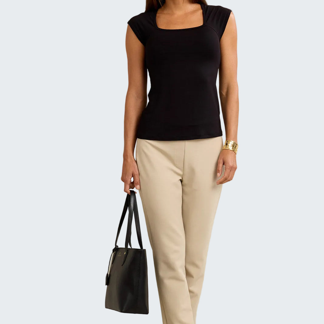 Woman wearing a black top and beige pants holding a black handbag against a neutral background