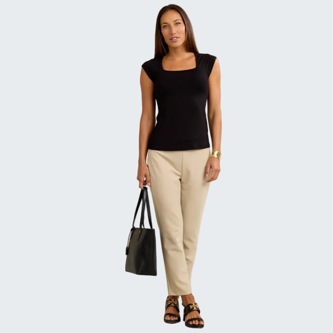 Woman wearing a black top and beige pants holding a black handbag against a neutral background