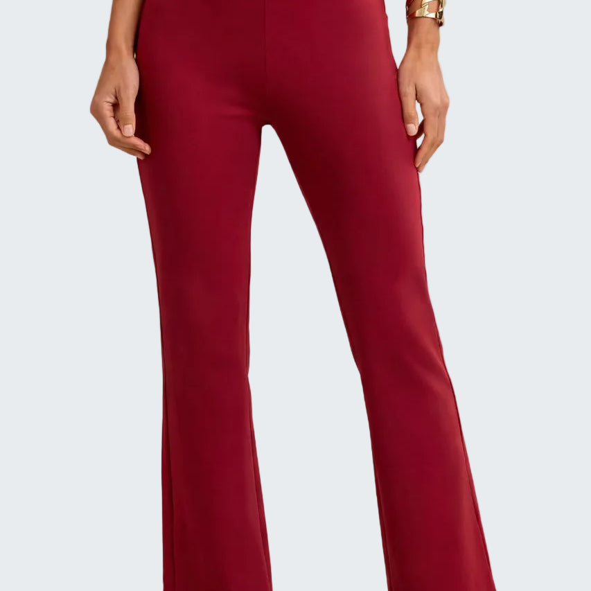 A model wearing high-waisted, straight-leg flared pants in a deep burgundy red, paired with patterned heeled sandals and a gold bracelet, against a neutral beige background.