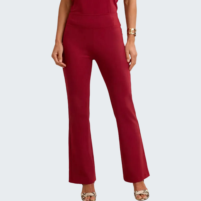 A model wearing high-waisted, straight-leg flared pants in a deep burgundy red, paired with patterned heeled sandals and a gold bracelet, against a neutral beige background.
