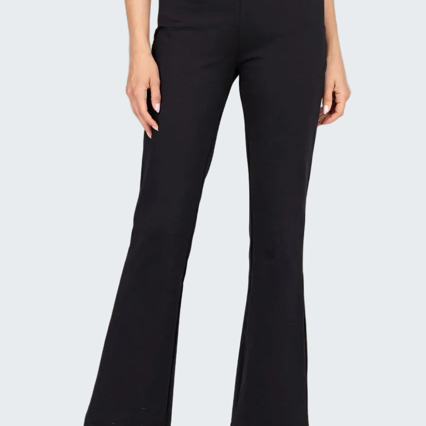 A model wearing black high-waisted flare pants with a smooth texture, paired with black sandals, against a neutral beige textured backdrop.