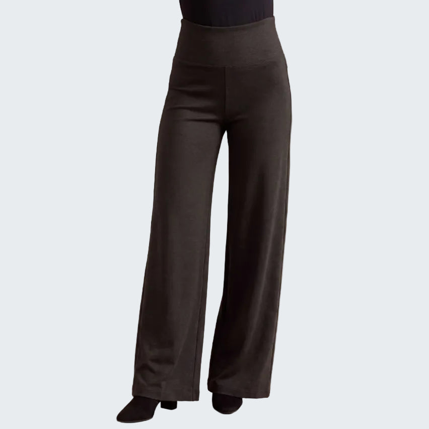 Black wide-leg pants shown on a model from the waist down, paired with a black top and black shoes, against a neutral beige textured background.