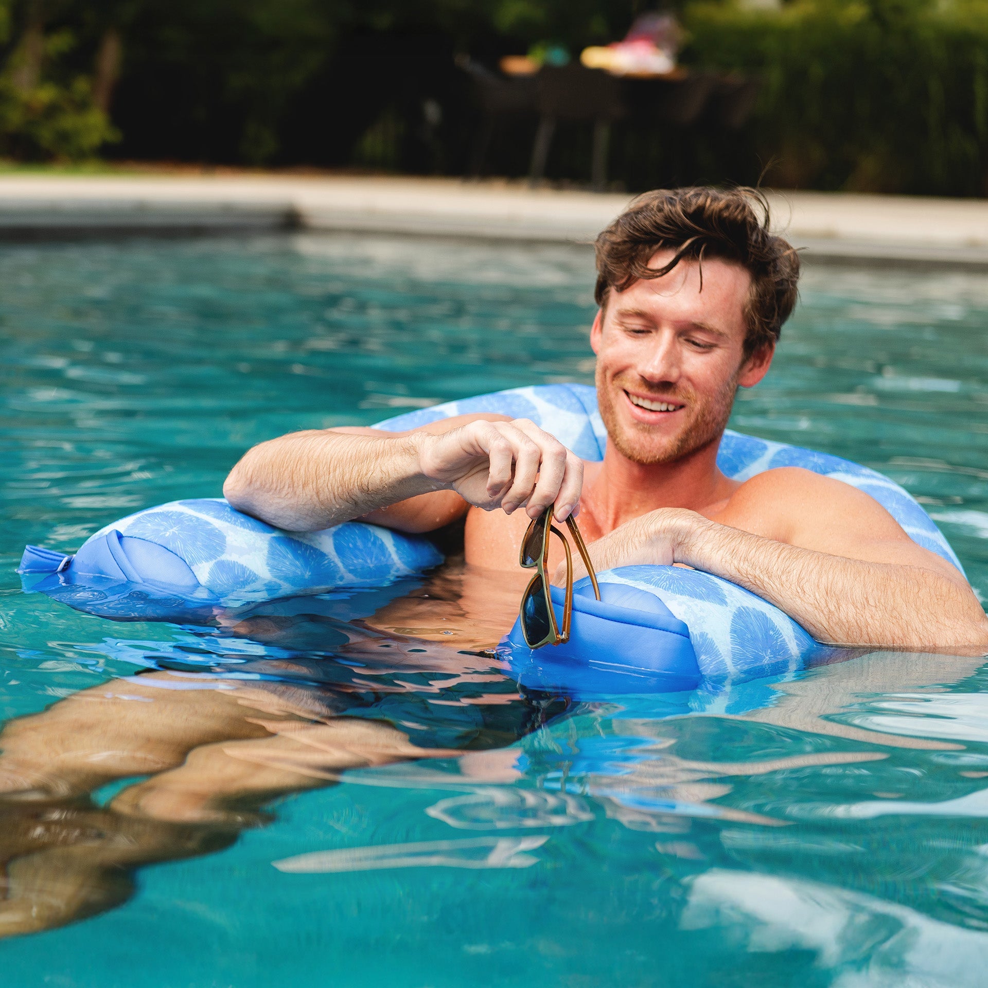 Man floating on a blue inflatable ring in a pool, holding sunglasses.