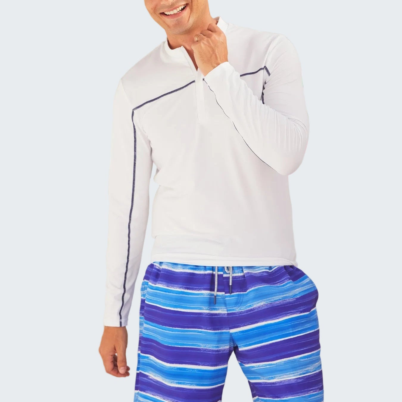 Man wearing a white long-sleeve shirt and blue striped shorts standing in front of a polka dot surfboard.
