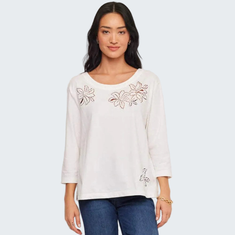 A woman modeling a white three-quarter sleeve top with embroidered floral details on the chest and lower hem, paired with blue jeans, against a plain light gray background.