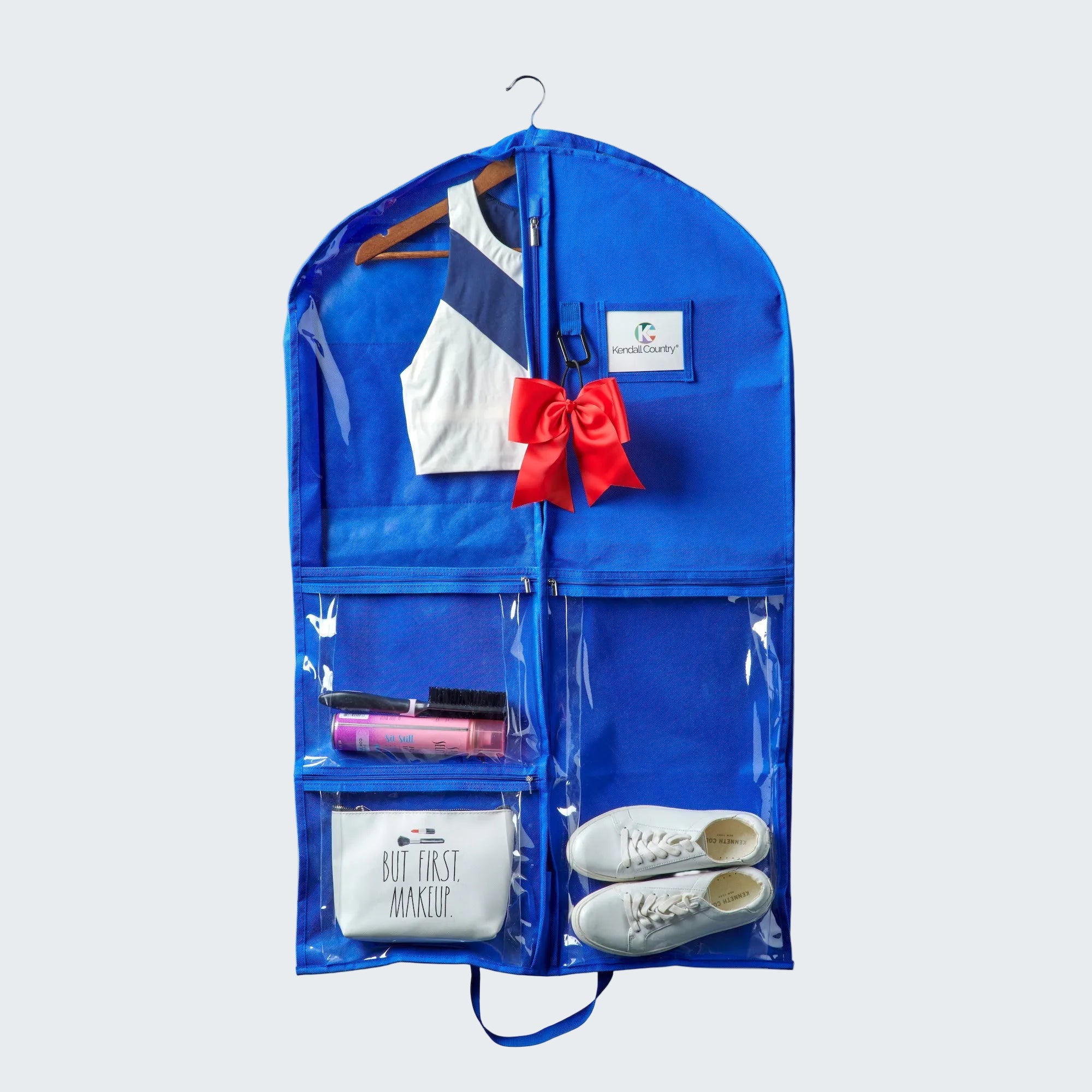 Blue garment bag with clear front pocket containing a white shirt, red bow, shoes, and makeup items on a white background.