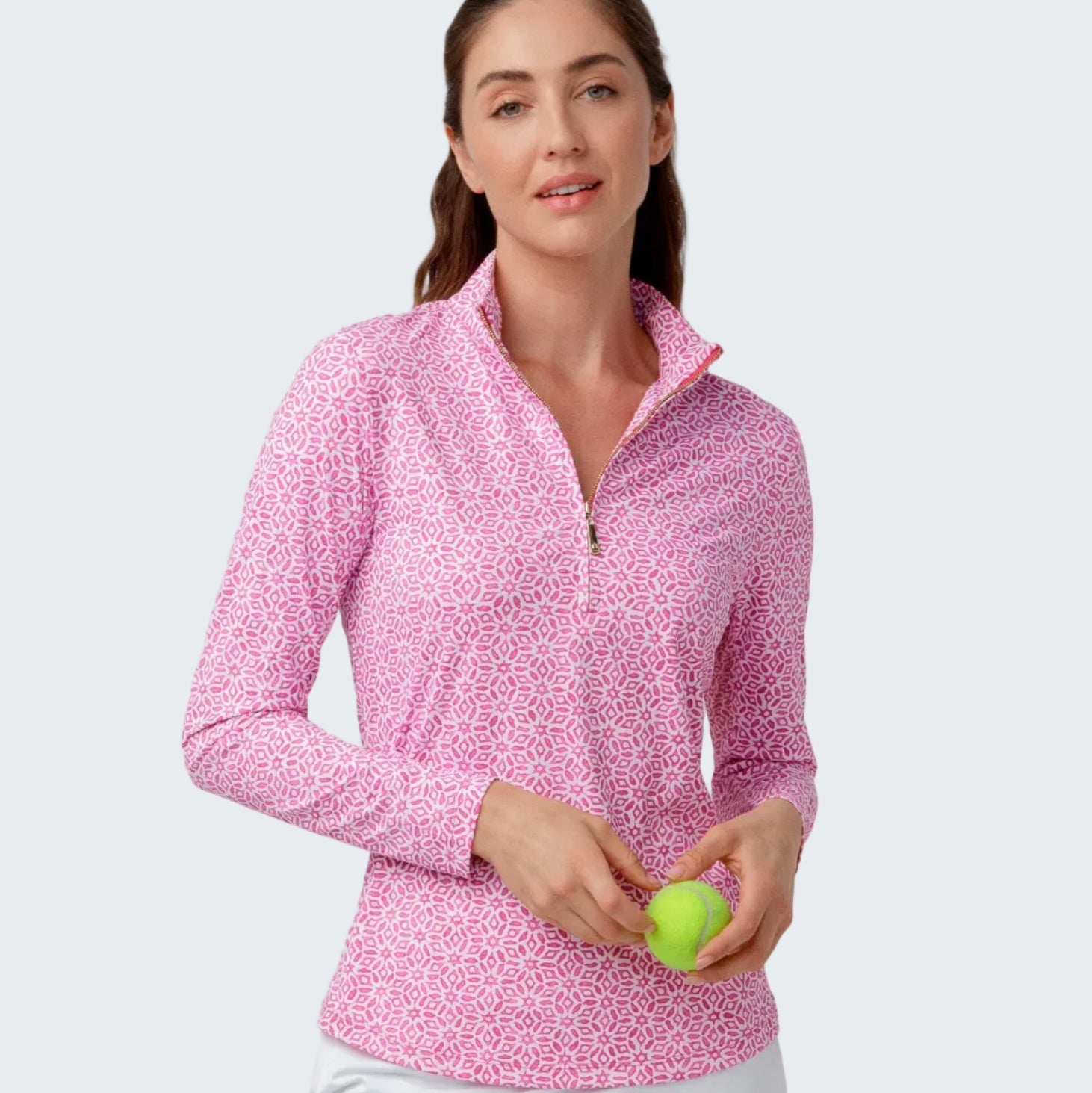 Woman wearing a pink patterned long-sleeve top and white golf skort, holding a green golf ball on a light gray background.