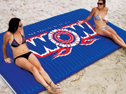 Two women sitting on a blue inflatable mat with 'WOW' branding on a sandy beach.