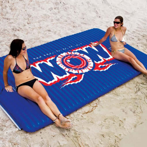 Two women sitting on a blue inflatable mat with 'WOW' branding on a sandy beach.