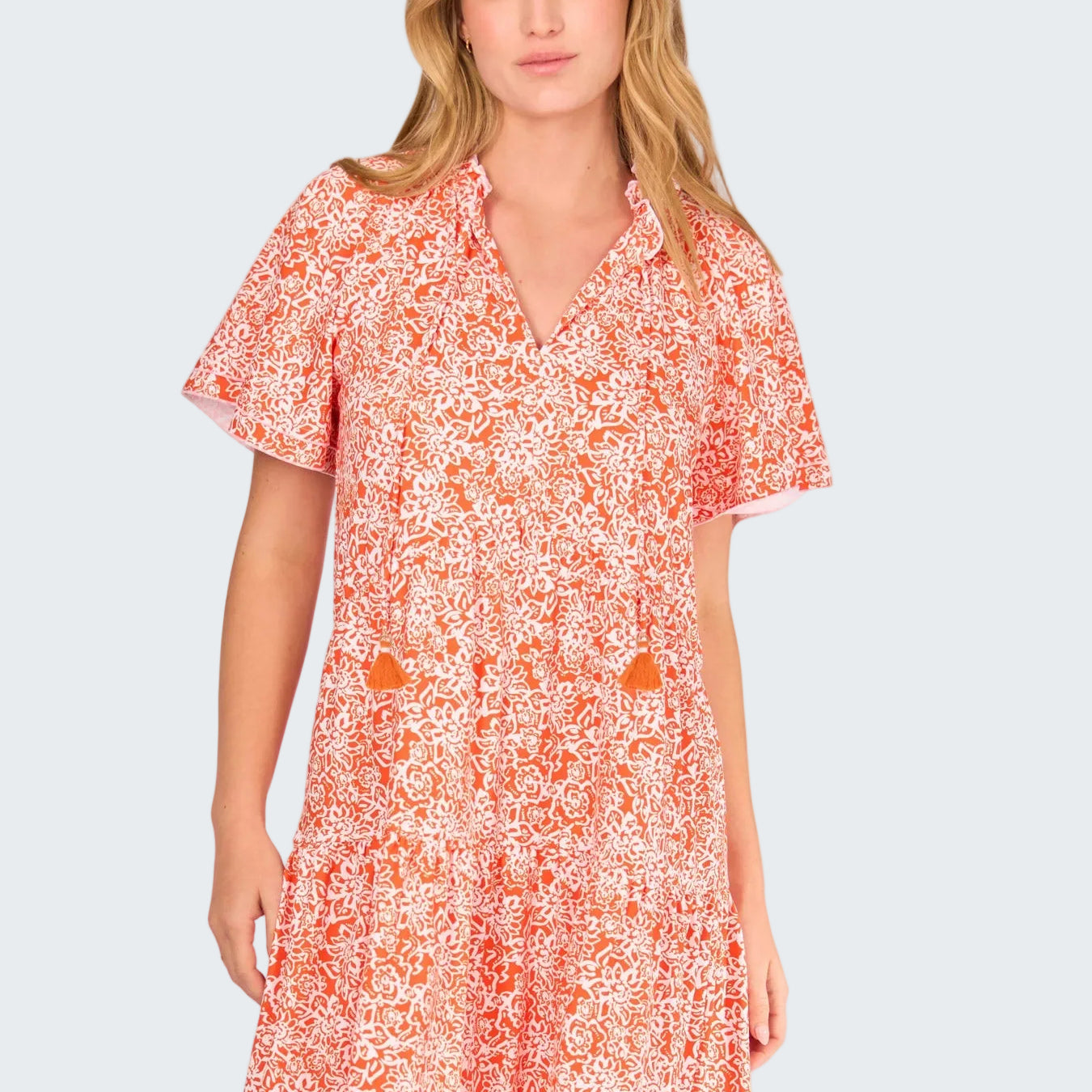A woman modeling a short-sleeve V-neck dress with a dense orange and white floral print against a solid white background.