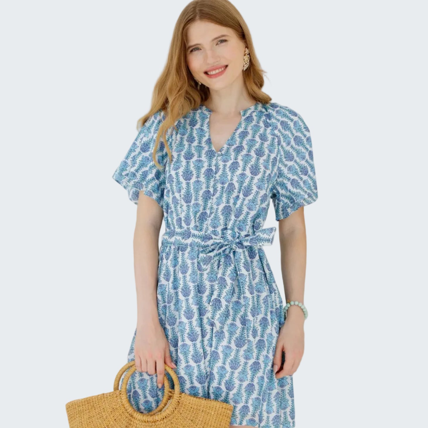 Woman wearing a blue patterned dress holding a straw bag against a white wall.