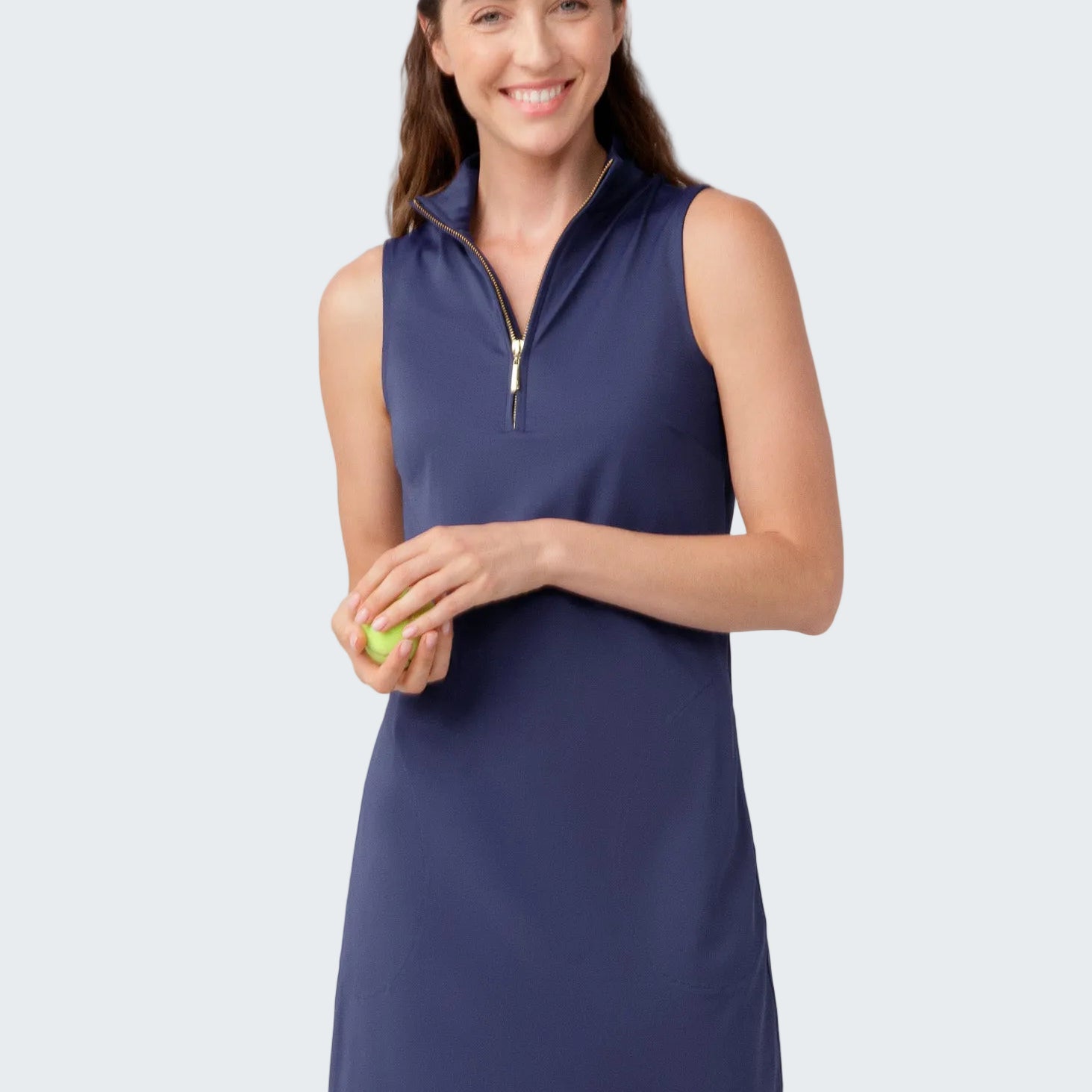 Woman wearing a navy sleeveless dress with a zipper, holding a tennis ball on a light gray background