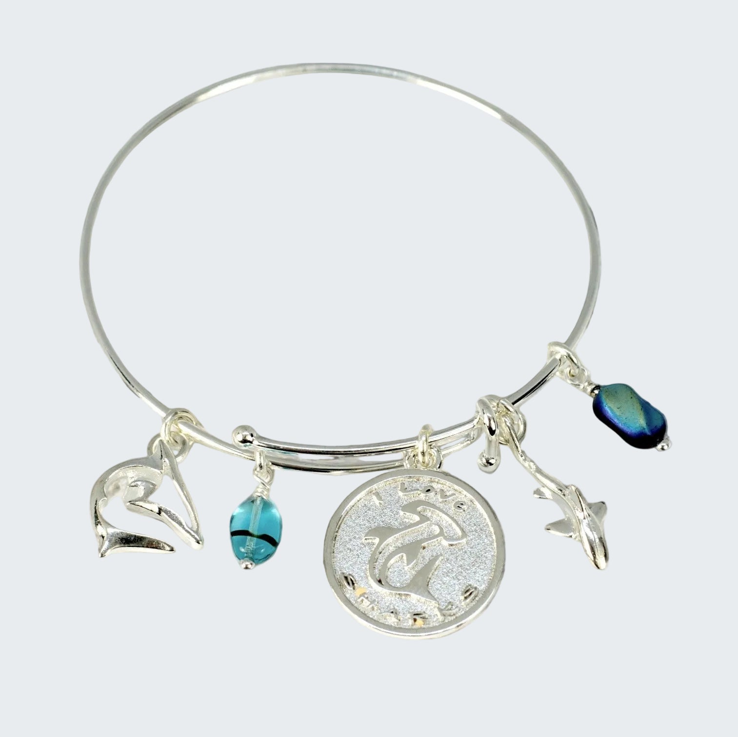 A silver bangle bracelet with sea-themed charms, including a dolphin pendant, a seashell charm, a starfish charm, and blue beads, displayed on a weathered wooden surface.