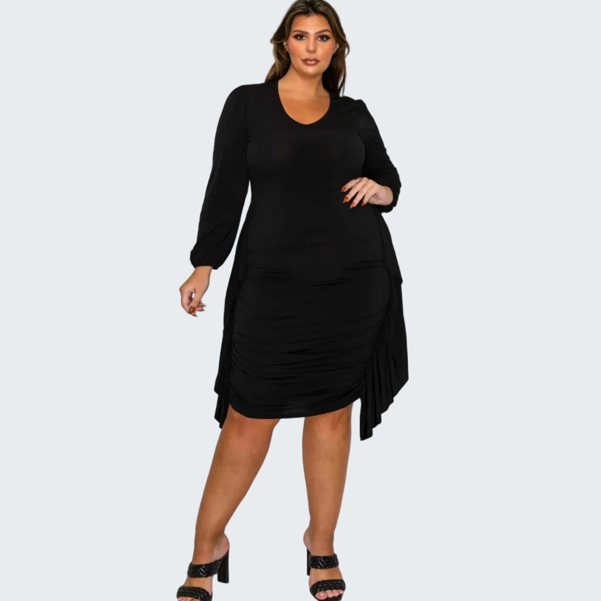 A woman modeling a black long-sleeve midi dress with ruched detailing and an asymmetrical draped hem, paired with black heeled sandals, against a plain white background.