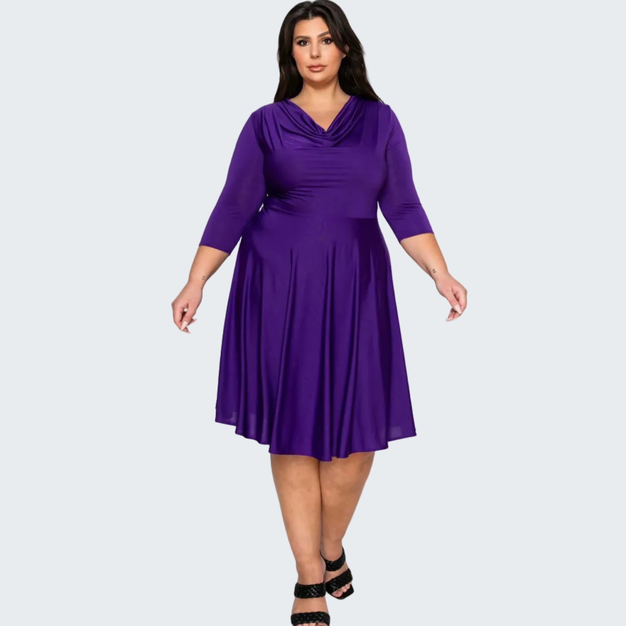 A woman modeling a purple plus-size cowl neck midi dress with three-quarter sleeves and a pleated skirt, paired with black strappy heels, against a solid white background.