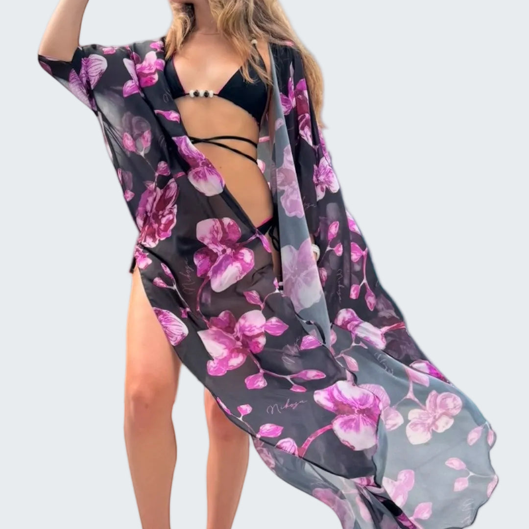 Woman in a floral kimono and black bikini standing on a rocky shore with ocean and sky in the background.