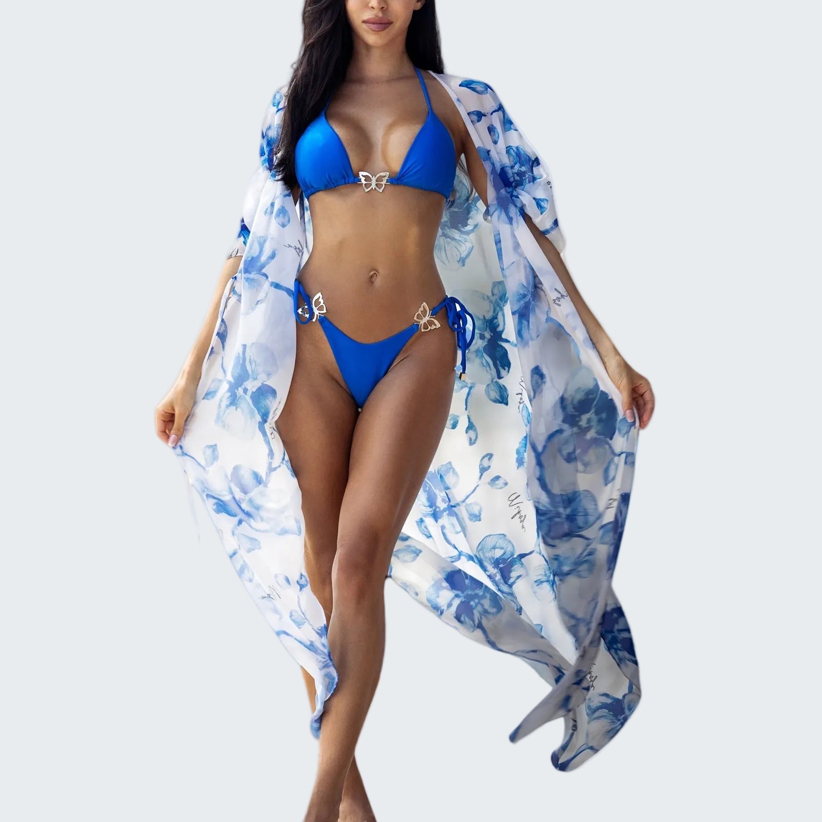 Woman in a blue bikini with a floral cover-up standing on a beach with ocean view.