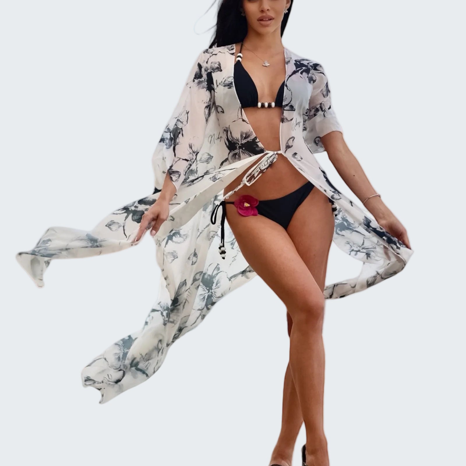 Woman in a black bikini with a floral cover-up standing on a stone ledge with a scenic background.