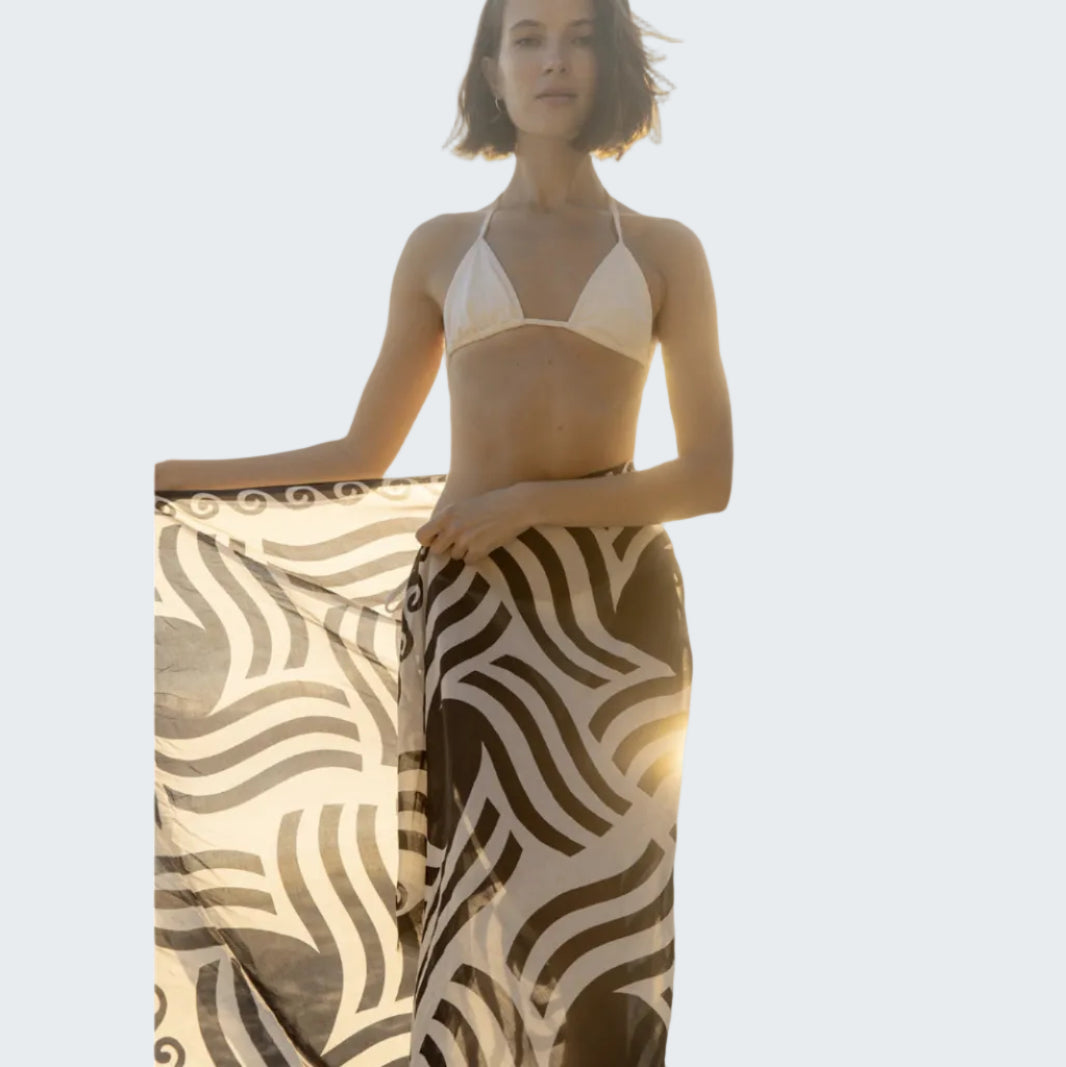 Woman in a bikini and patterned sarong standing on a beach.