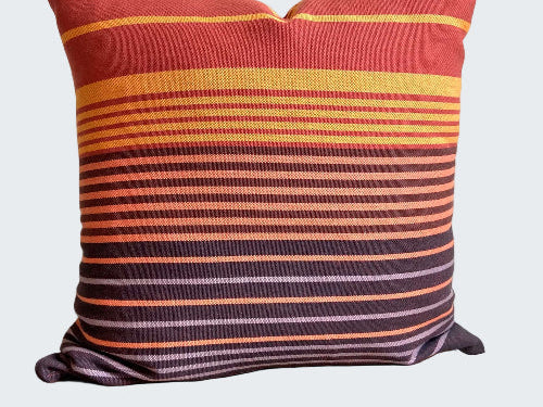 Striped pillow with red, orange, and purple colors on a wooden stool against a white wall.