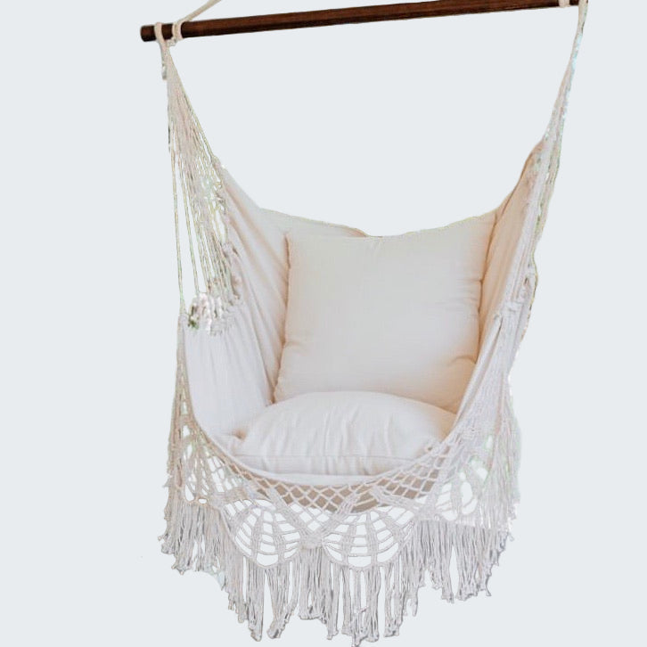 Macrame hammock chair with cushions on a wooden deck, surrounded by greenery.