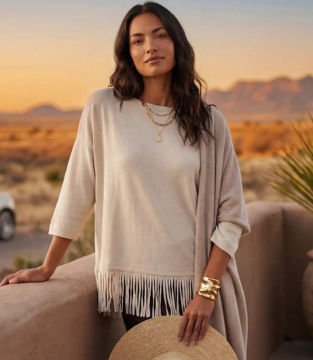 A woman wearing a cream-colored knit top with fringe trim and a matching draped cardigan, accessorized with gold jewelry, standing outdoors in a desert landscape at sunset.
