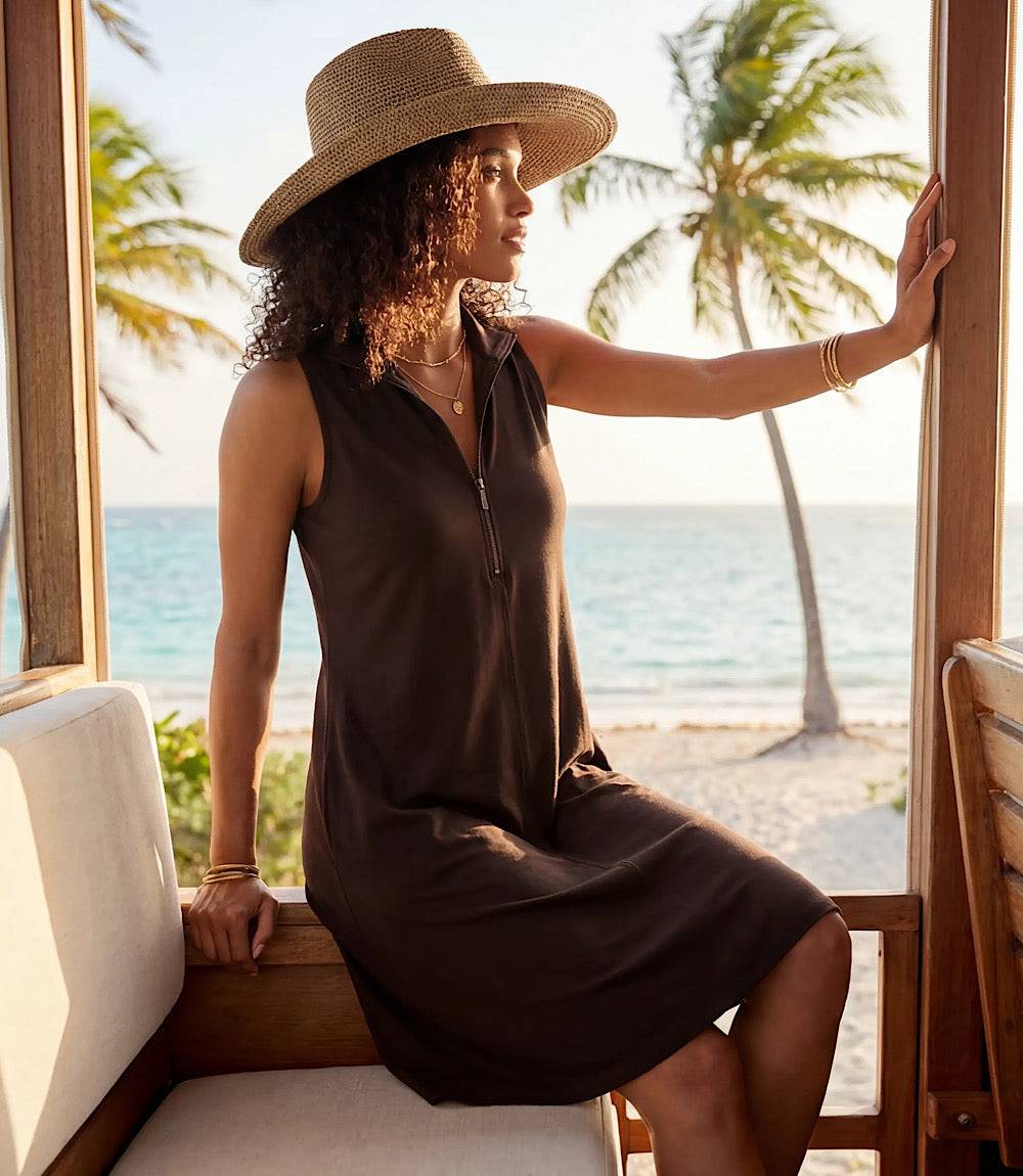 Woman in a brown dress and hat sitting by a window with a beach view