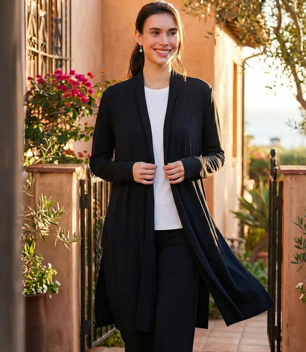A woman stands outdoors wearing a long black open-front cardigan over a white top and black pants. The setting has warm stucco walls, plants, and a gate, with sunlight and a blurred background.