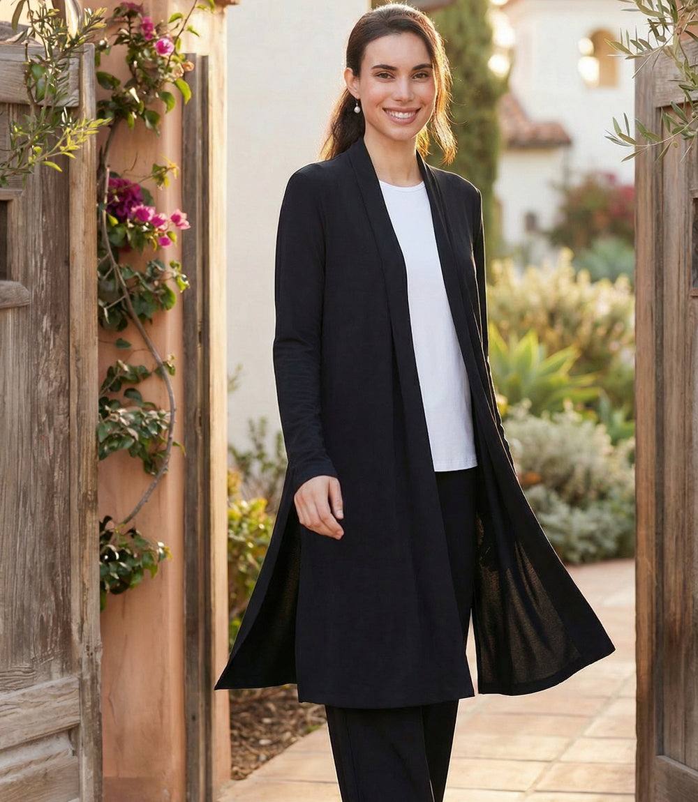 A woman stands in a garden doorway wearing a long black open-front cardigan over a white top and black pants. The background shows warm-toned wooden doors, climbing flowers, and soft-focus greenery.