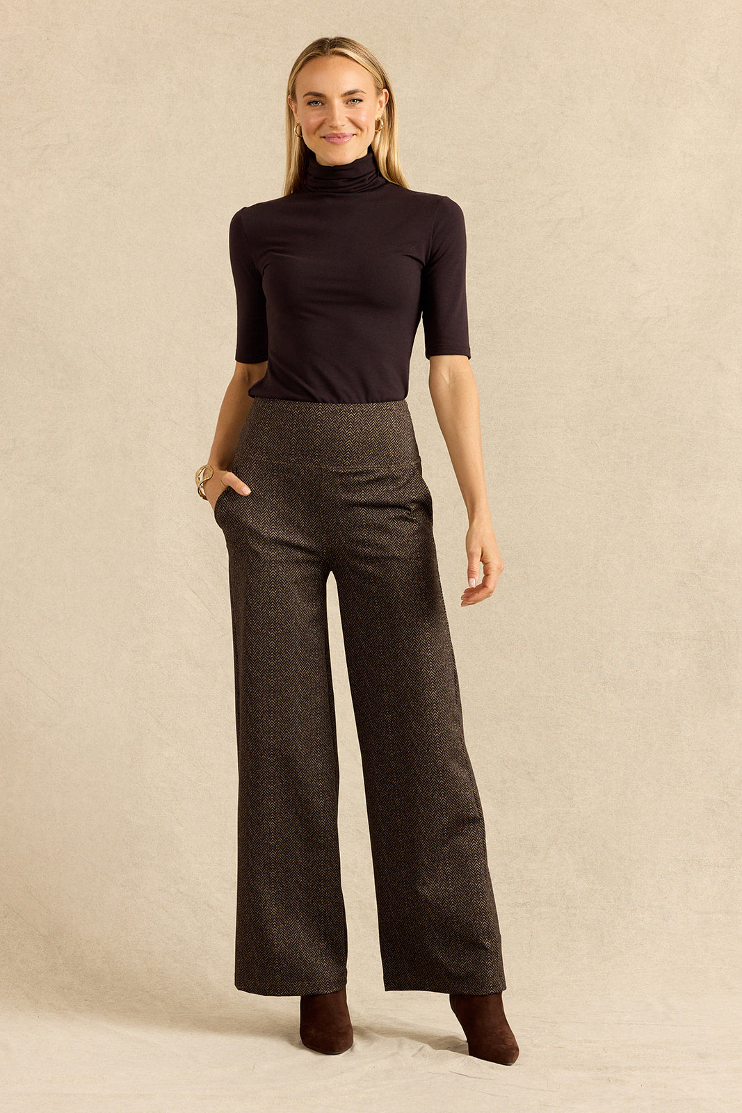 A woman modeling a dark brown heathered wide-leg trouser set with a matching dark turtleneck top, standing against a textured beige studio backdrop.