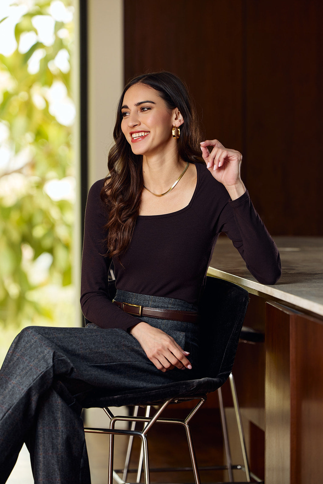 A woman with long dark wavy hair sits on a black bar stool in a modern interior, wearing a dark plum long-sleeve scoop-neck top, dark gray wide-leg jeans, and a brown belt.