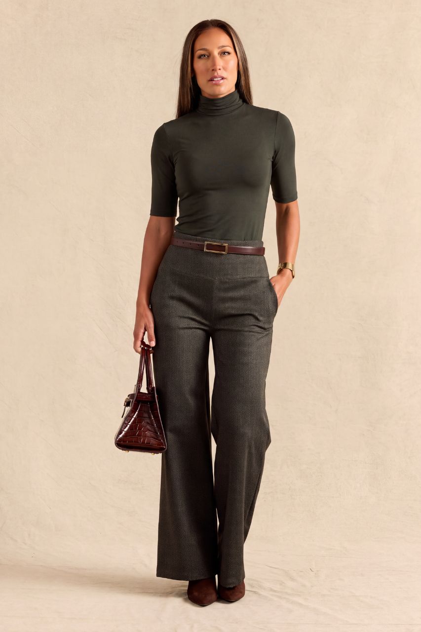 A woman modeling a dark olive green short-sleeve turtleneck top and matching dark gray wide-leg trousers, styled with a brown belt, brown shoes, and a dark brown handbag against a textured beige studio backdrop.