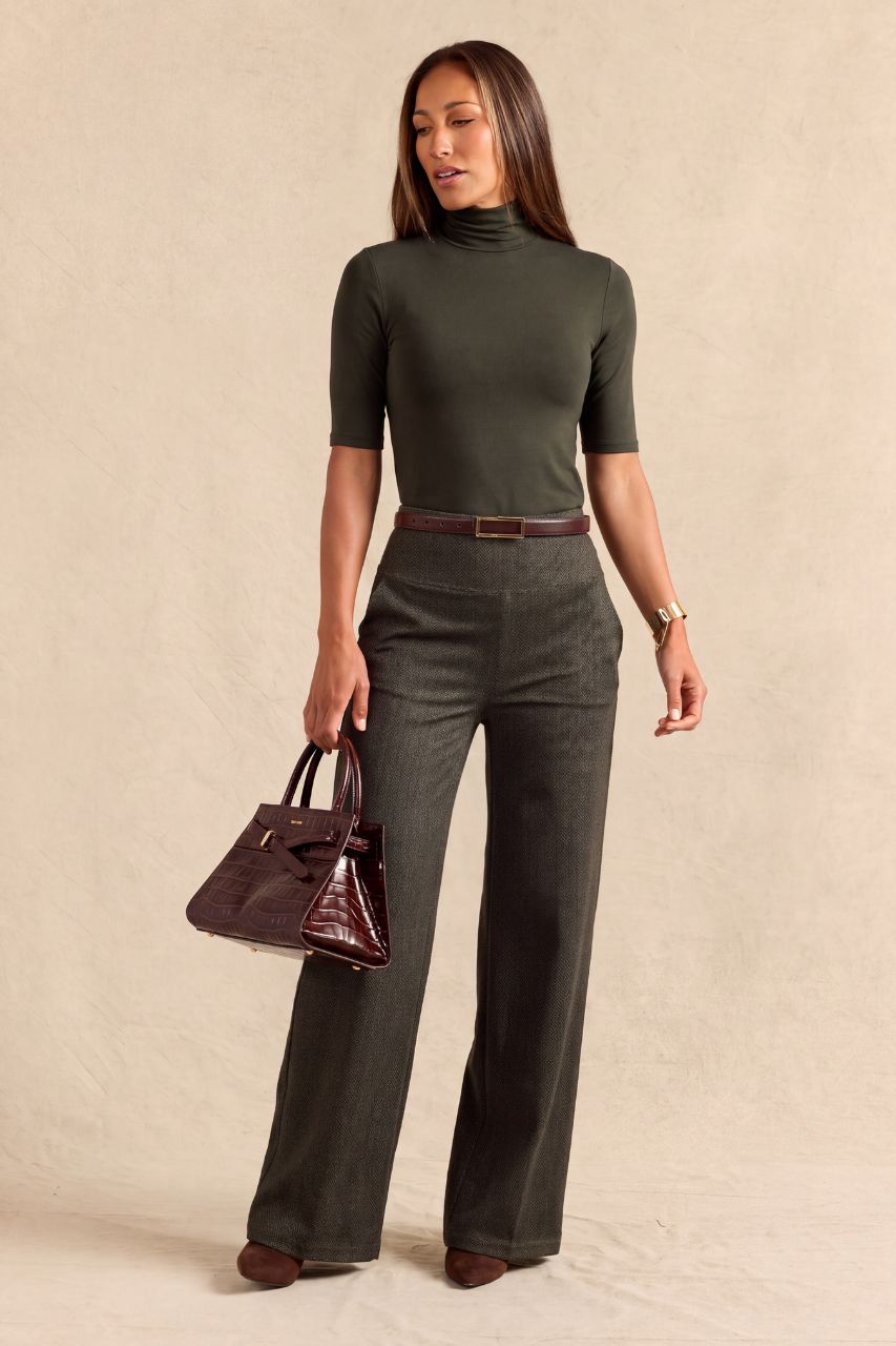 A woman modeling an olive green short-sleeve mock neck top and matching dark olive green wide-leg trousers, styled with a brown belt, brown shoes, and a dark burgundy handbag against a textured beige studio backdrop.