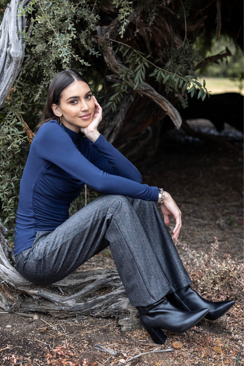 A woman sits outdoors against a large tree trunk, wearing a fitted navy long-sleeve top, wide-leg charcoal gray pants, and black ankle boots.
