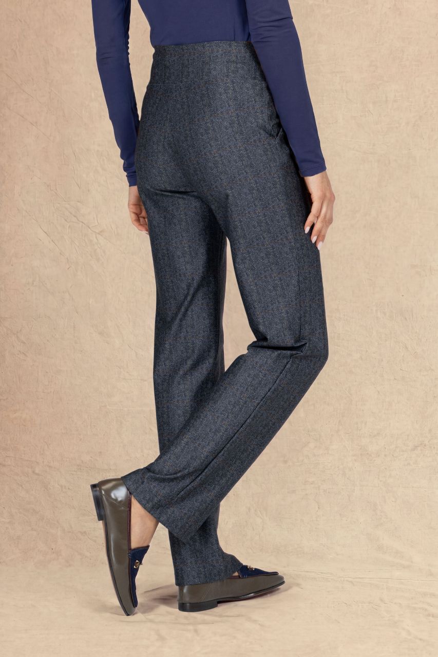 A model from the back wearing high-waisted, wide-leg charcoal gray tweed trousers, a navy blue long-sleeve top, and dark loafers, standing against a textured beige backdrop.