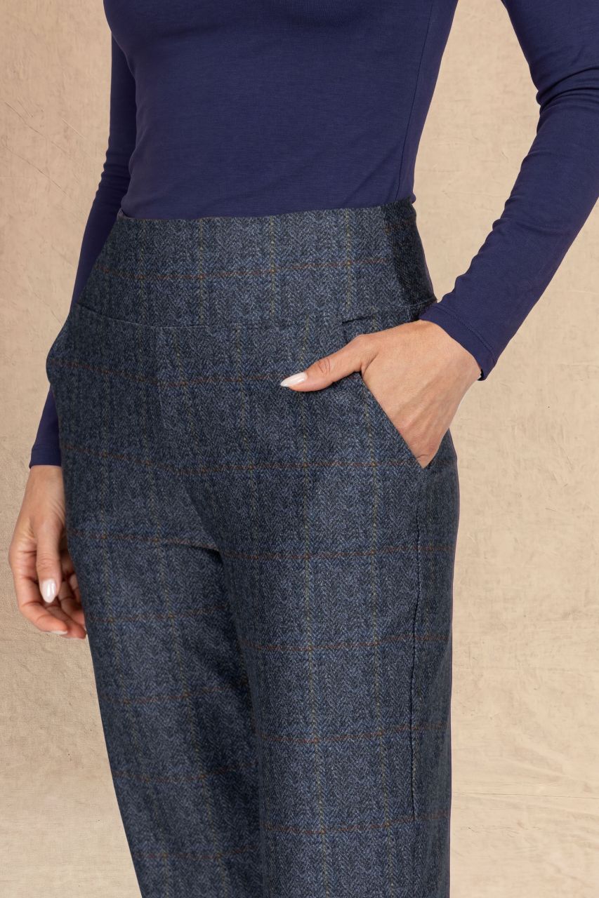 A close-up of a person wearing high-waisted navy blue windowpane plaid trousers and a navy long-sleeve top, standing against a textured beige backdrop.