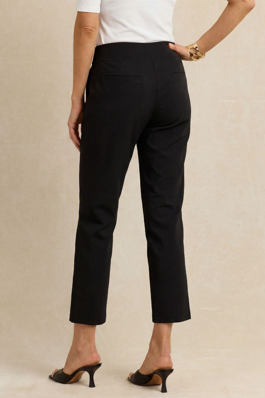 Rear view of a model wearing black cropped trousers with a white top and black mule heels, standing against a neutral beige background.