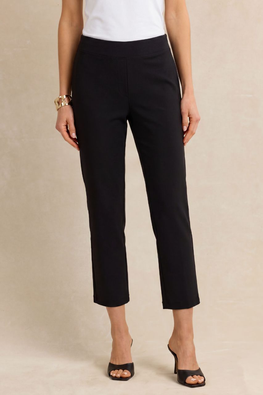 A model wearing black cropped ankle-length trousers, paired with a white top and black heeled sandals, standing against a neutral beige background.