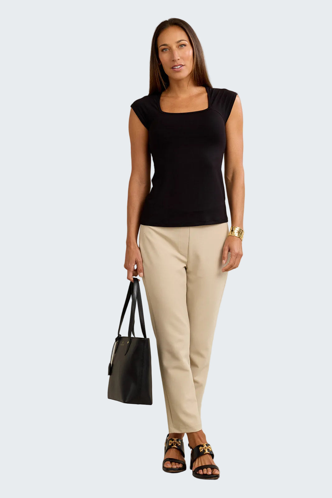 Woman wearing a black top and beige pants holding a black handbag against a neutral background