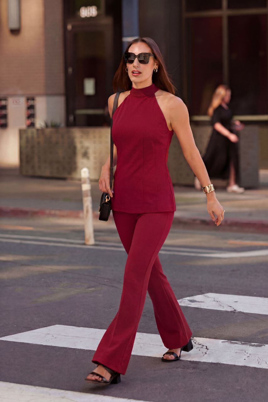 A woman walking on a city crosswalk wearing a matching sleeveless high-neck top and flared pants set in deep burgundy, accessorized with black sunglasses, a black shoulder bag, and black sandals.