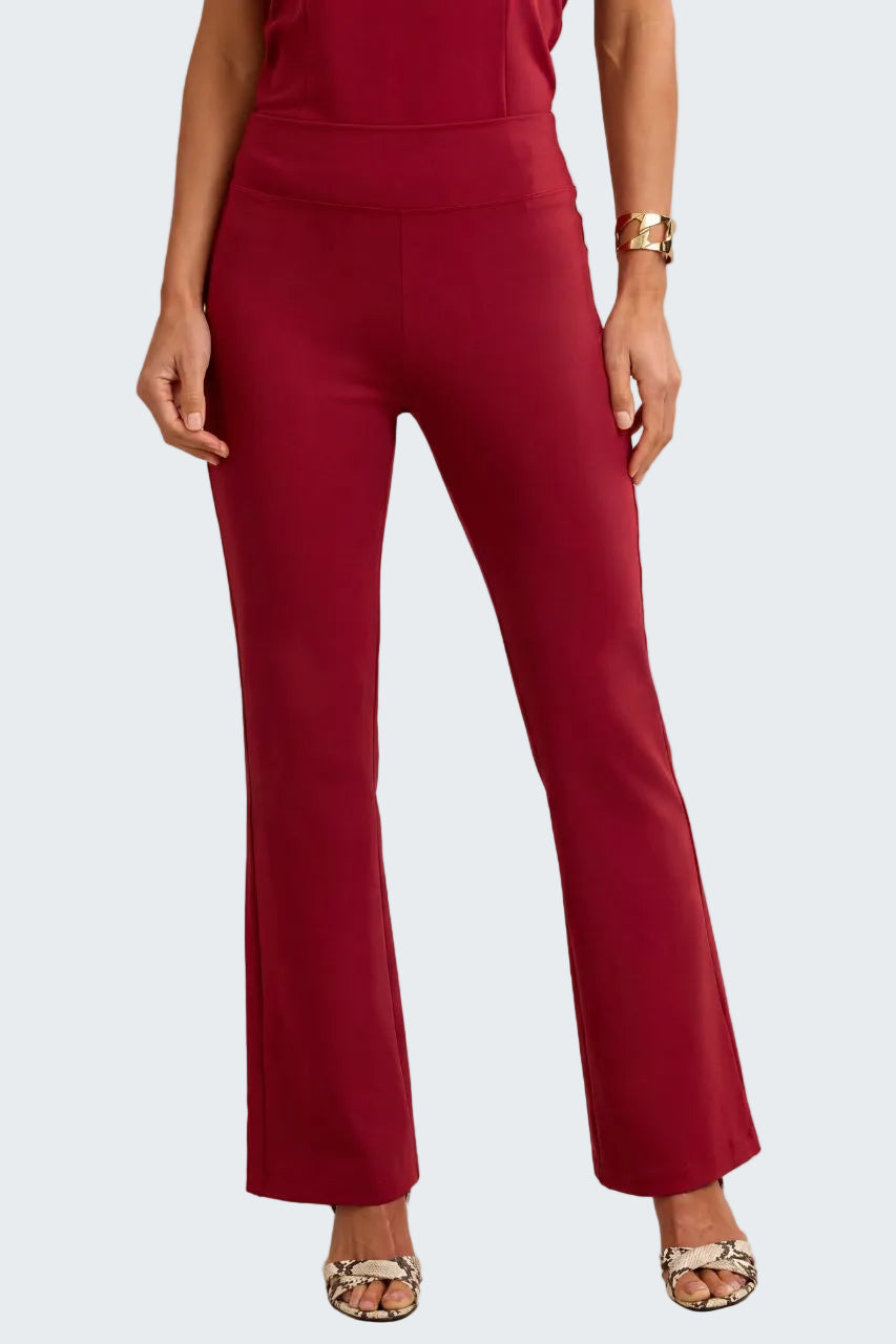 A model wearing high-waisted, straight-leg flared pants in a deep burgundy red, paired with patterned heeled sandals and a gold bracelet, against a neutral beige background.
