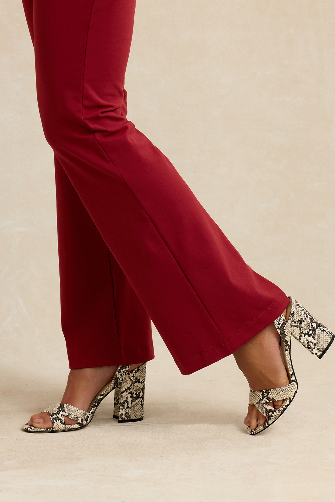 A close-up of a person's legs wearing deep red flared trousers and beige-and-black snakeskin-patterned block-heel sandals against a warm beige studio background.