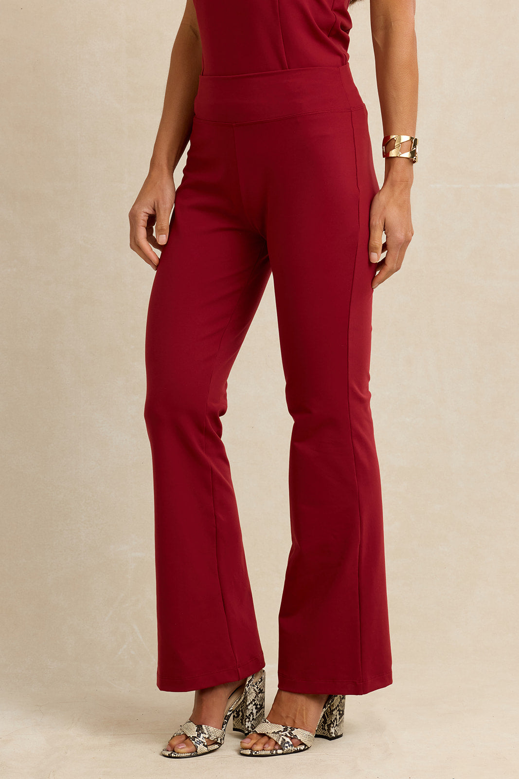 A model is shown from the waist down wearing high-waisted, wide-leg flared trousers in a deep burgundy red. The pants are paired with snakeskin-patterned heeled sandals and a gold bracelet, set against a plain beige studio background.