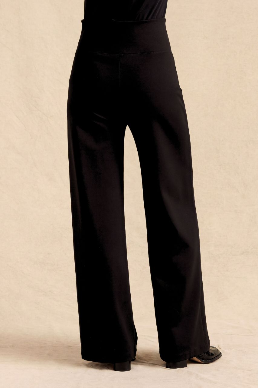 Black wide-leg trousers shown from the back on a model, featuring a high-waisted design and a smooth satin-like finish, set against a neutral beige textured background.
