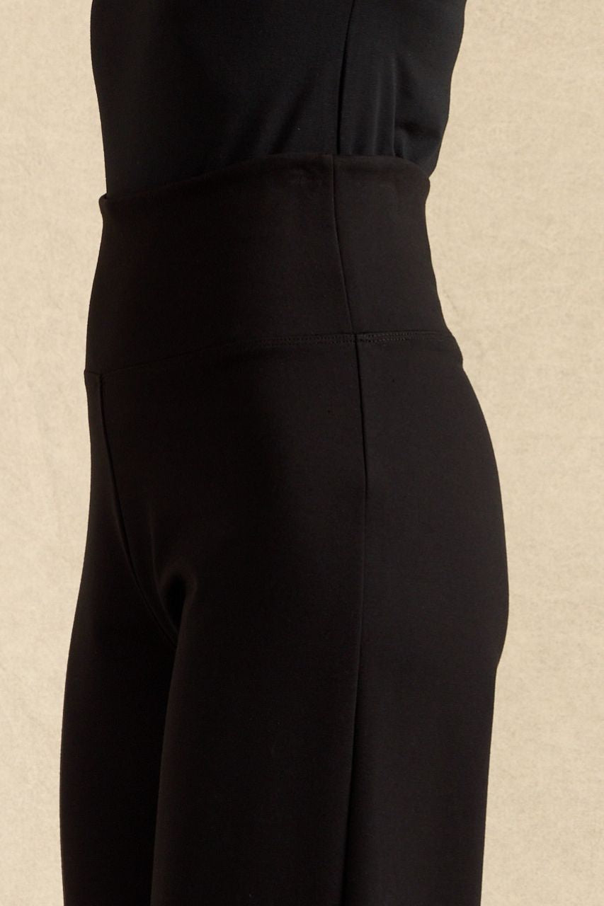 Close-up side view of a person wearing black high-waisted cropped pants against a warm beige textured background.