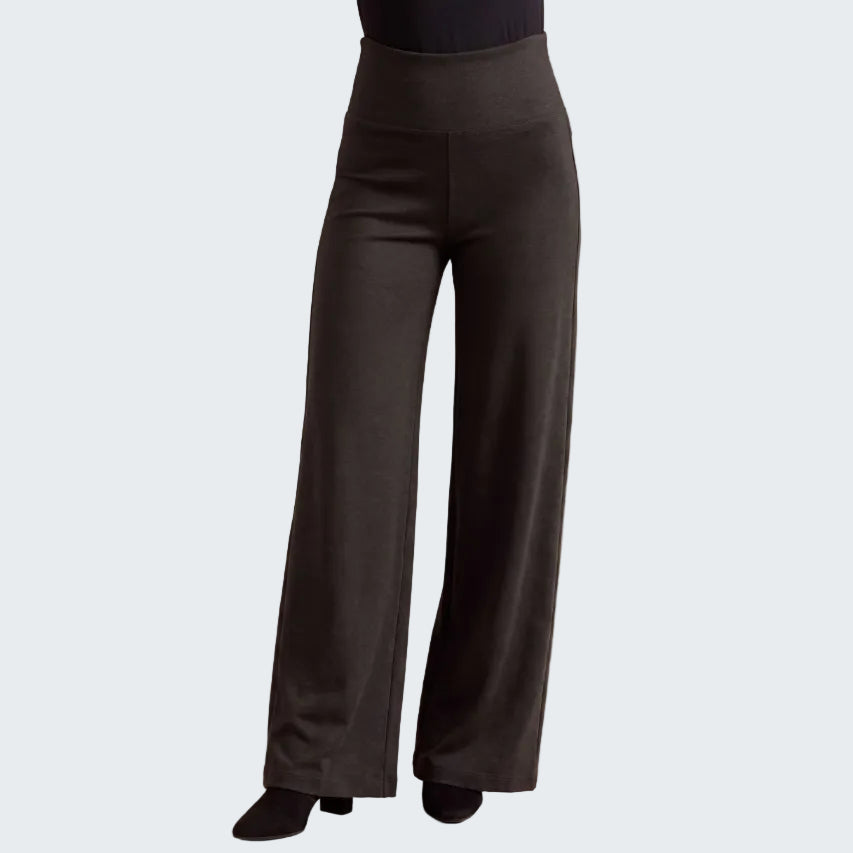 Black wide-leg pants shown on a model from the waist down, paired with a black top and black shoes, against a neutral beige textured background.