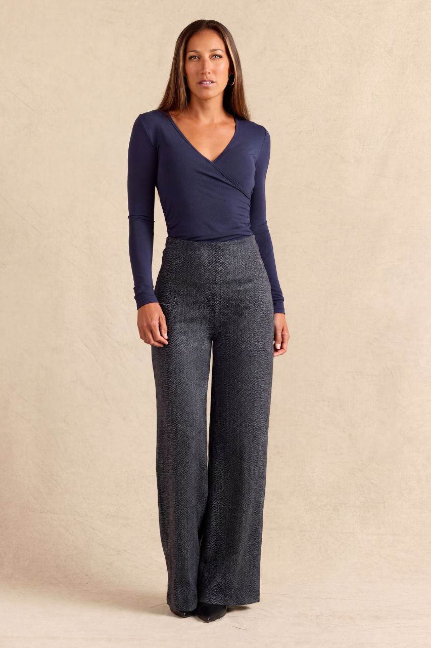 A woman modeling a navy blue long-sleeve wrap top and dark gray wide-leg pants against a textured beige studio backdrop.