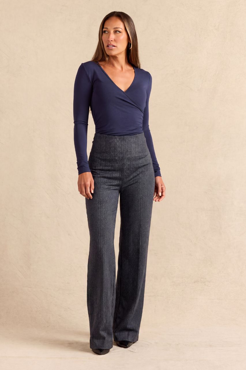 A woman modeling a navy blue long-sleeve wrap-style top paired with dark gray wide-leg pants, standing against a textured beige studio backdrop.