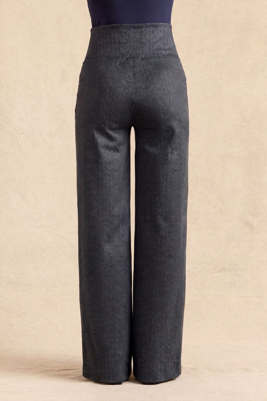 A pair of dark gray wide-leg trousers shown from the back on a model, with a navy top partially visible at the top edge, set against a textured beige backdrop.