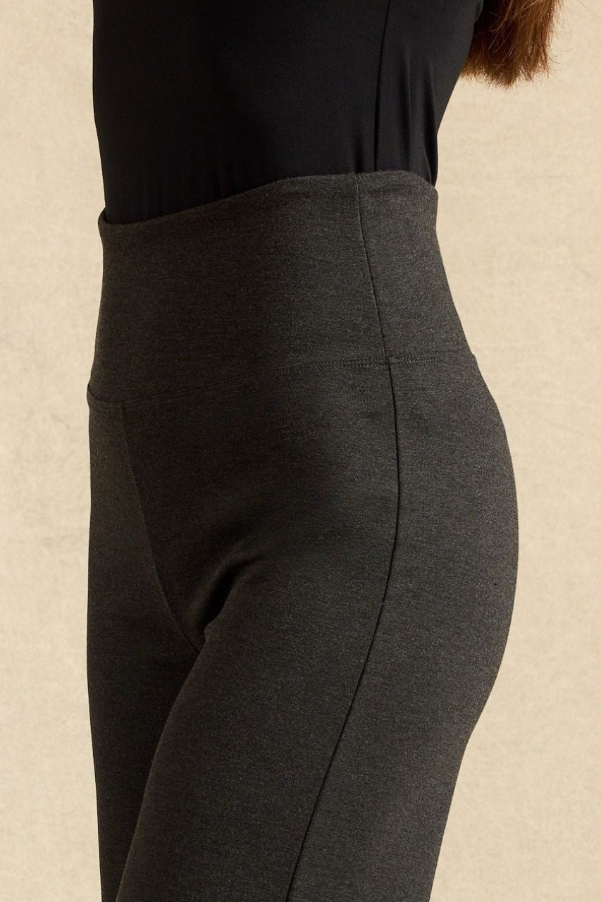 Close-up side view of a person wearing dark charcoal gray high-waisted leggings with a black top, against a neutral beige background.